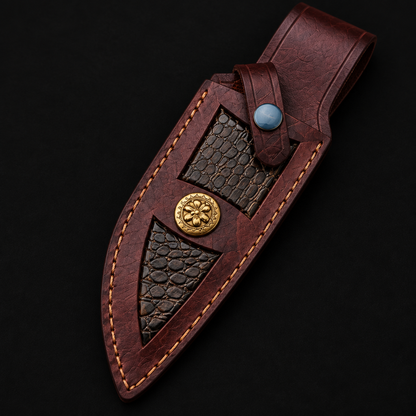 Extreme close-up of premium burgundy leather hunting knife sheath with snake skin inlay panels and gold floral medallion snap button on black background