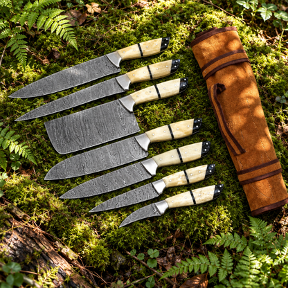 Handmade 7-piece Damascus steel chef knife set cream bone handles and tan suede leather roll bag on thick green forest moss natural outdoor
