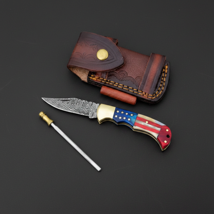 Handmade Damascus folding pocket knife with American flag resin handle, leather sheath, and sharpening rod on black-charcoal background