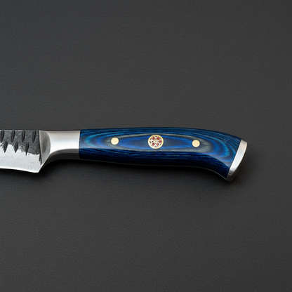 Close view of the blue stained pakkawood handle with brass pins, brass mosaic pin, and stainless steel end cap on black charcoal