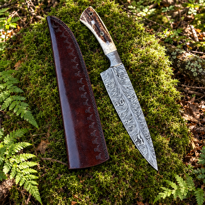 Handmade feather Damascus chef knife antler handle and oxblood tooled leather sheath on thick green forest moss natural outdoor
