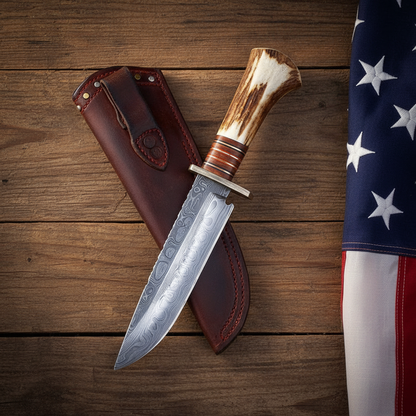Handmade Damascus Bowie hunting knife with stag antler handle and dark brown leather sheath on rustic wood with an American flag