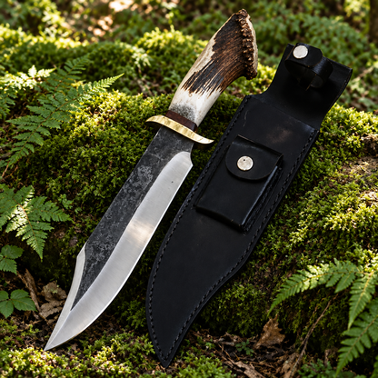 Carbon steel brut de forge Bowie knife and black leather sheath resting on thick green forest moss natural outdoor setting
